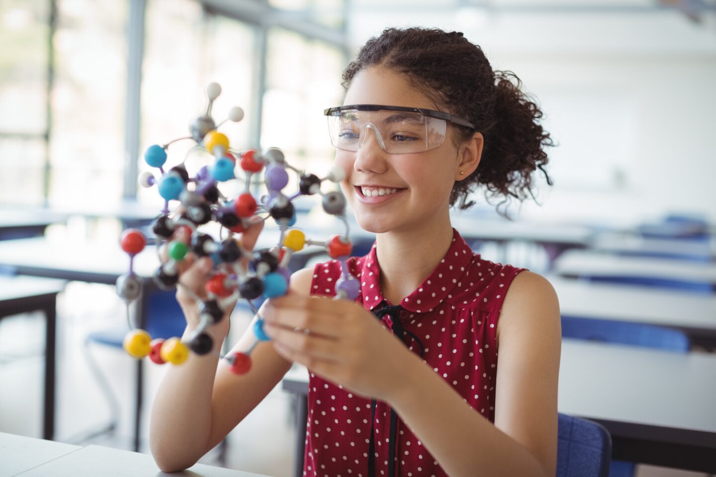 Girl in her safety goggles looking at an atom structure smiling, studying fr multiple science A Level exam boards.  